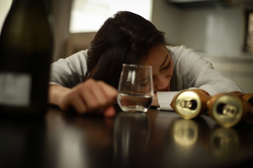 woman lays on table surrounded by a half full glass and empty cans and bottles suffering from symptoms of alcohol poisoning